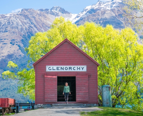 Lady standing in doorway of red Glenorchy wharf shed in front of mountains and green trees