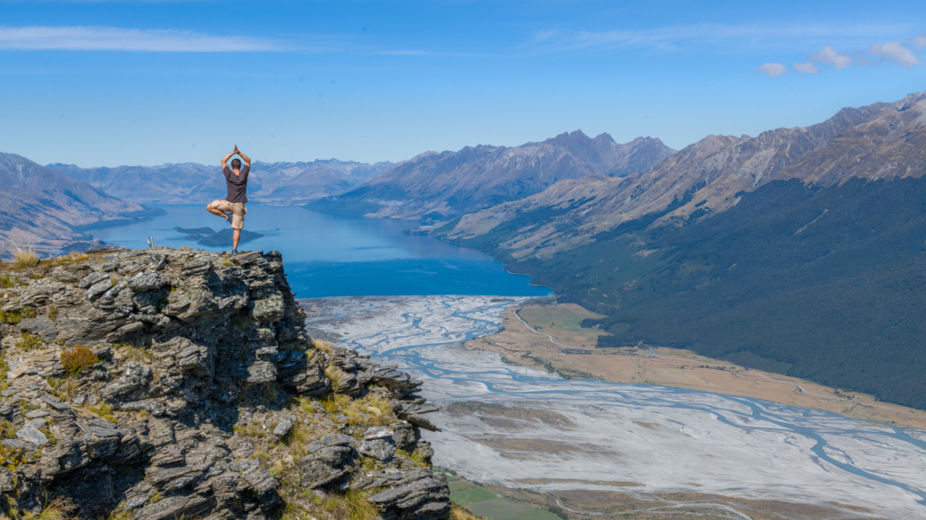 Man doing yoga pose on top of Mt Alfred with view of Lake Wakatipu, mountains and Kinloch