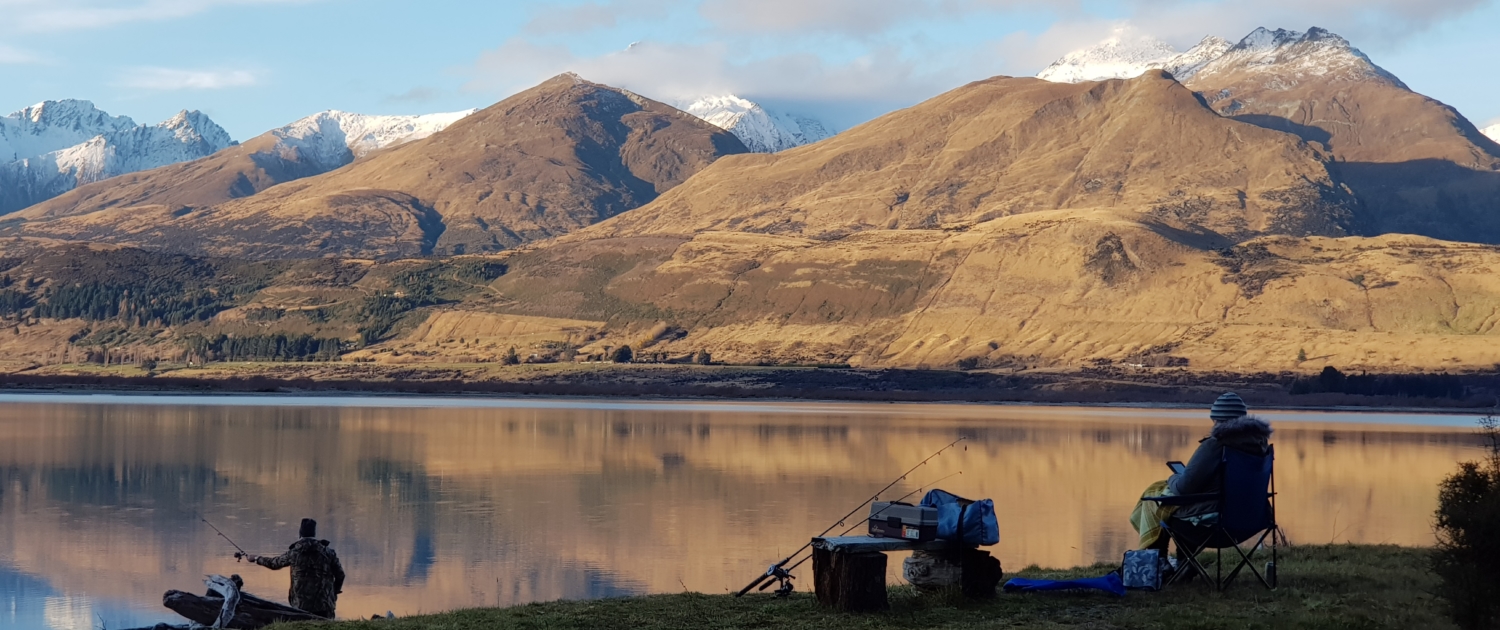 Man fishing Lake Wakatipu in evening and lady sat reading with mountain view