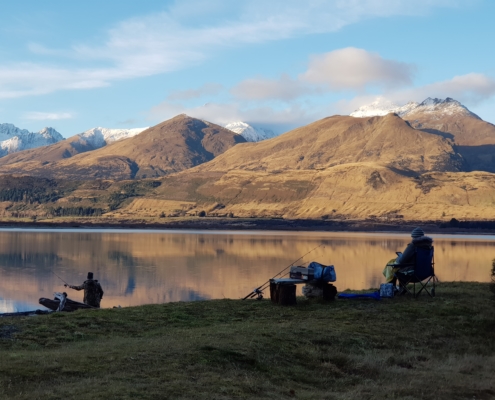Man fishing Lake Wakatipu in evening and lady sat reading with mountain view