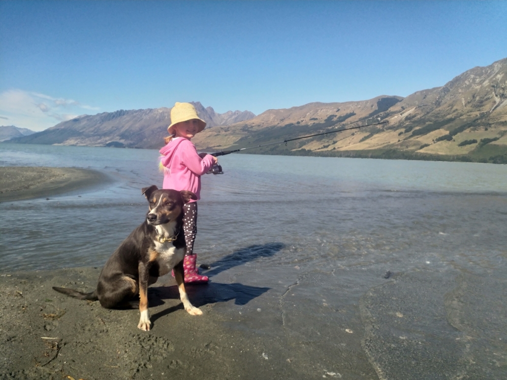 Girl fishing on the edge of Lake Wakatipu with a dog sitting next to her